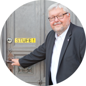 Thomas Domres steht vor einer Haustür mit dem Schild "Stufe!", die Hand liegt auf der Türklinke